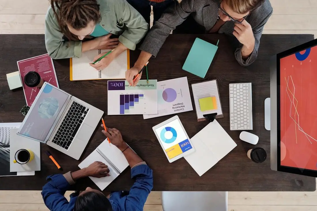 Four people collaborate at a table covered with laptops, notes, and charts, discussing data analysis and strategy in a modern workspace.