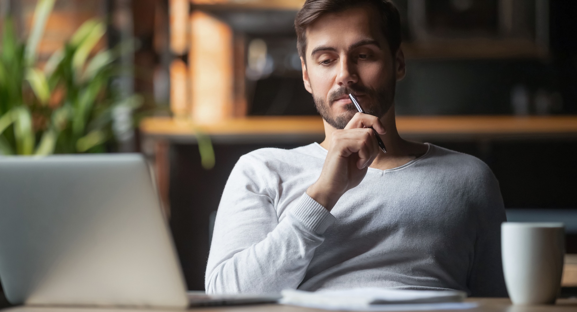 A thoughtful man sits at a desk with a laptop, pen in hand, contemplating ideas. A coffee mug and notepad are nearby, suggesting productivity.